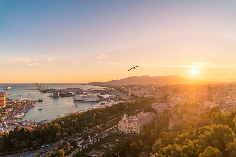 Zračni zalazak sunca iznad luke, marine i panorame Malage, sa vidljivim tornjem katedrale, točkom za vožnju i pristanim trajektima duž obale dok galeb prolijeće.