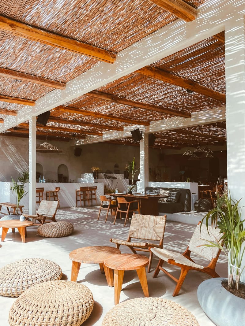 Lounge na otvorenom beach kluba s pletenim ratan stolicama, okruglim drvenim stočićima, podnim pufovima i pergolom od trske koja baca prugaste sjene.