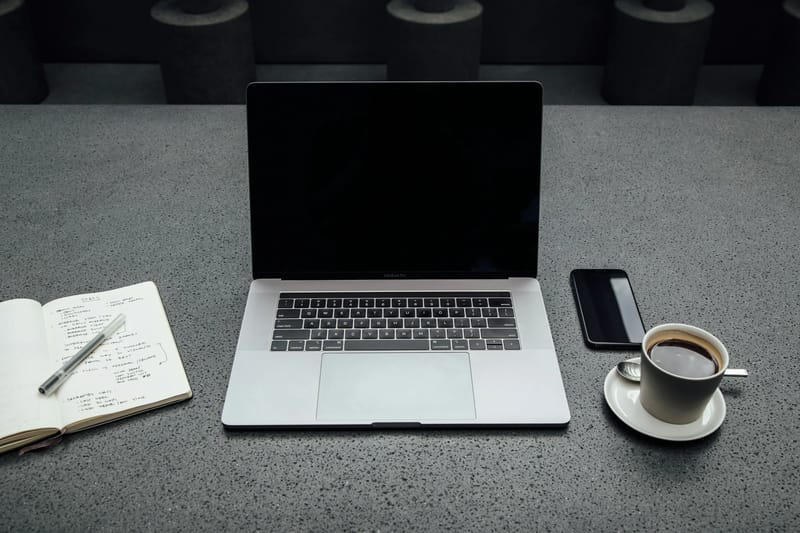 Një MacBook Pro me ekran të mbyllur qëndron mbi një tavolinë betoni midis një blloku shënimesh të hapur me stilolaps, një iPhone dhe një filxhan kafeje në një nënnytë.
