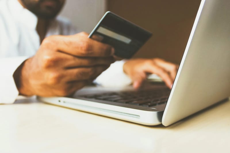 Një burrë mban një kartë krediti me njërën dorë ndërsa shtyp në tastierën e laptopit me tjetrën, duke sugjeruar një pagesë ose rezervim online.