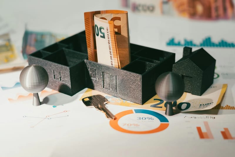 Mali 3D model kuće i stabala stoje na otisnutim finansijskim grafikonima i euro banknotama, s ključem od kuće pored, ilustrujući koncept ulaganja u nekretnine ili hipoteke.