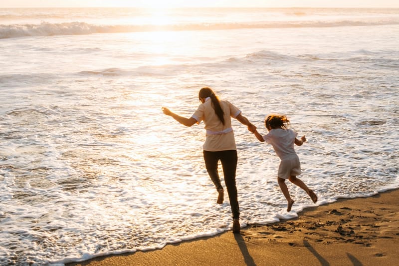 Majka i malo dijete, prikazani od pozadi, drže se za ruke i trče prema plitkom moru na plaži dok sunce zalazi, bacajući dugo zlatno svjetlo i sjene preko mokrog pijeska.
