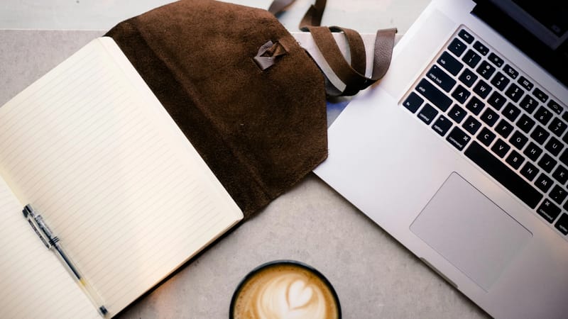 Otvoreni prazni rokovnik s olovkom, smeđom torbom od brušene kože, rubom srebrnog laptopa i šoljom kafe s latte art srcem poredani su na betonskom stolu.