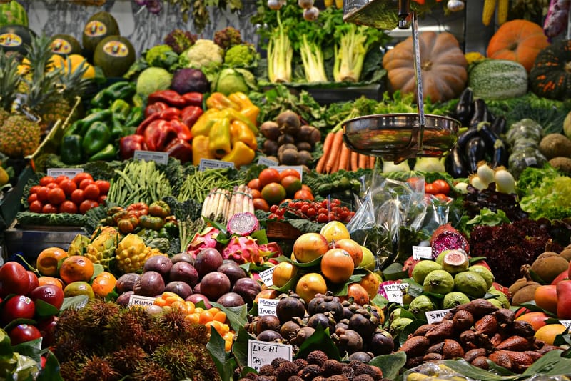 Një stendë e ngjeshur në një treg prodhimesh spanjolle me fruta dhe perime ekzotike (lichi, kumquat, granadilla, guayaba, salak) të etiketuara me shenja çmimesh të shkruara me dorë, me një peshore bronzi të varur mbi to.