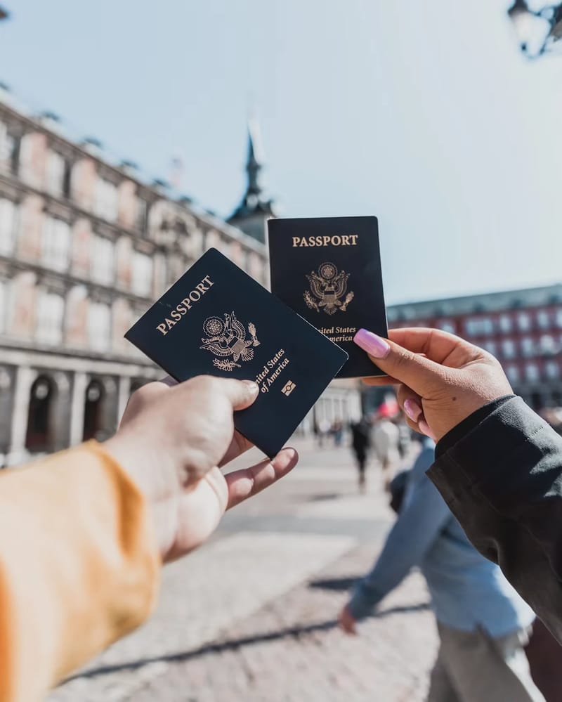 Dvoje ruku drži američke pasoše ispred istorijskog evropskog trga s ukrašenom ciglanom sat tornjem i prolaznicima u pozadini.